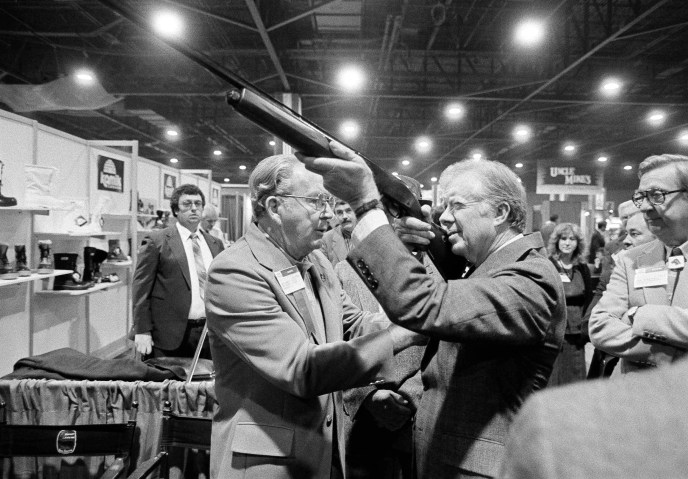 Former President Jimmy Carter sights down the barrel of a shotgun as Reinhart Fajen, a gunstock manufacturer, checks the fit, in Atlanta as Carter stopped during a visit to the national shooting sports foundation shot show at the World Congress Center on Nov. 14, 1984.